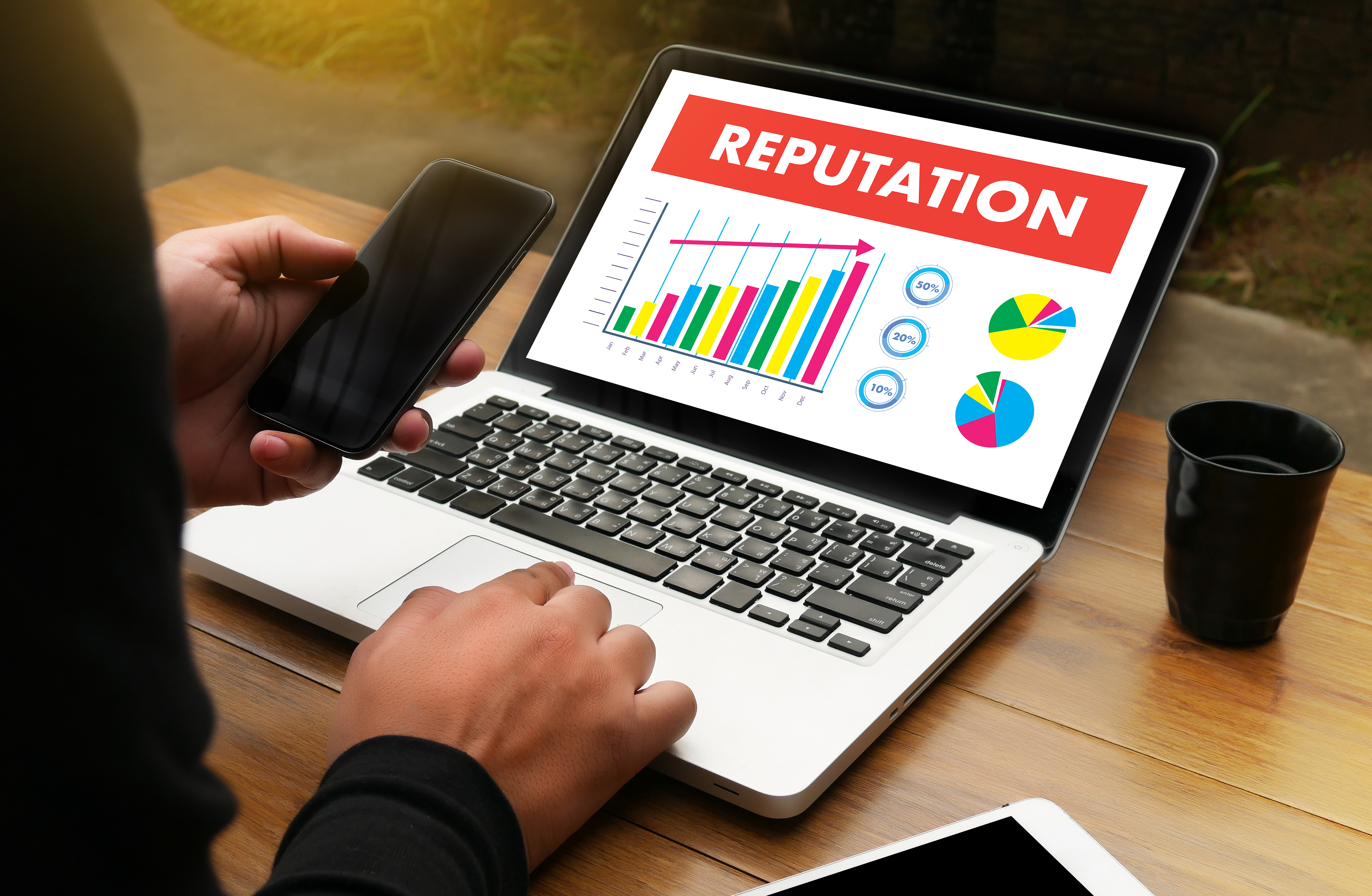 A person sitting a desk, you can't see their face. In their left hand, the hold a mobile phone, and with their right hand they navigae a laptop. On the laptop screen, a slide with the word Repuation lines the top, and multiple graphs are at the bottom of the screen.